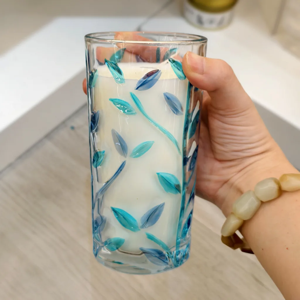 Hand holding a tall glass candle jar decorated with raised blue and teal leaf patterns.
