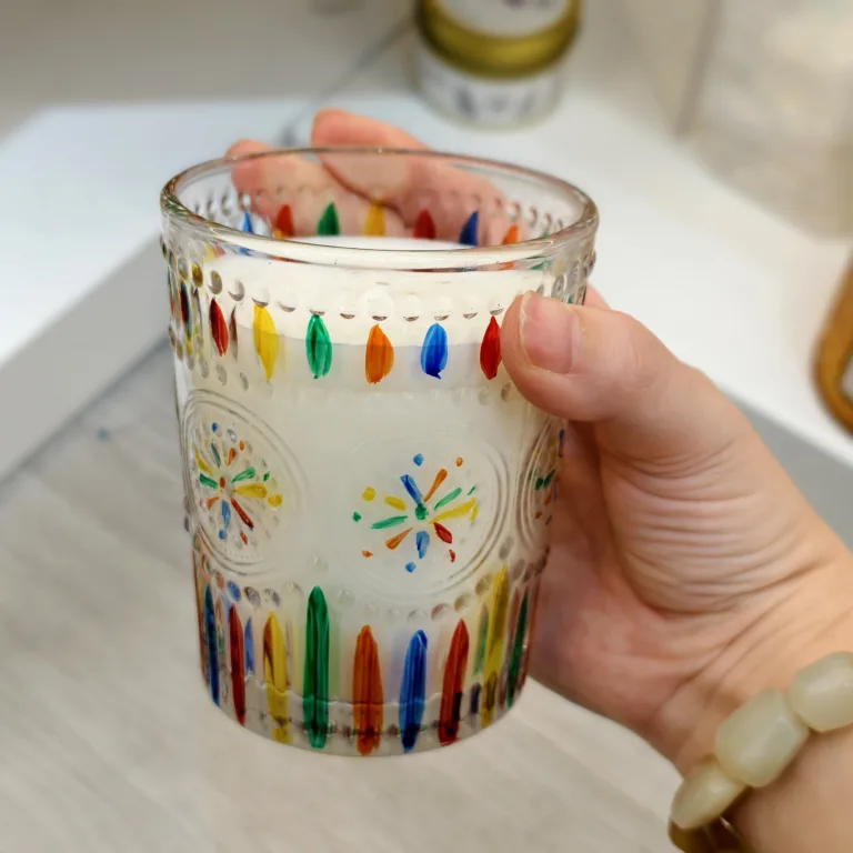 Hand holding a round glass candle jar with colorful painted dots and starburst patterns